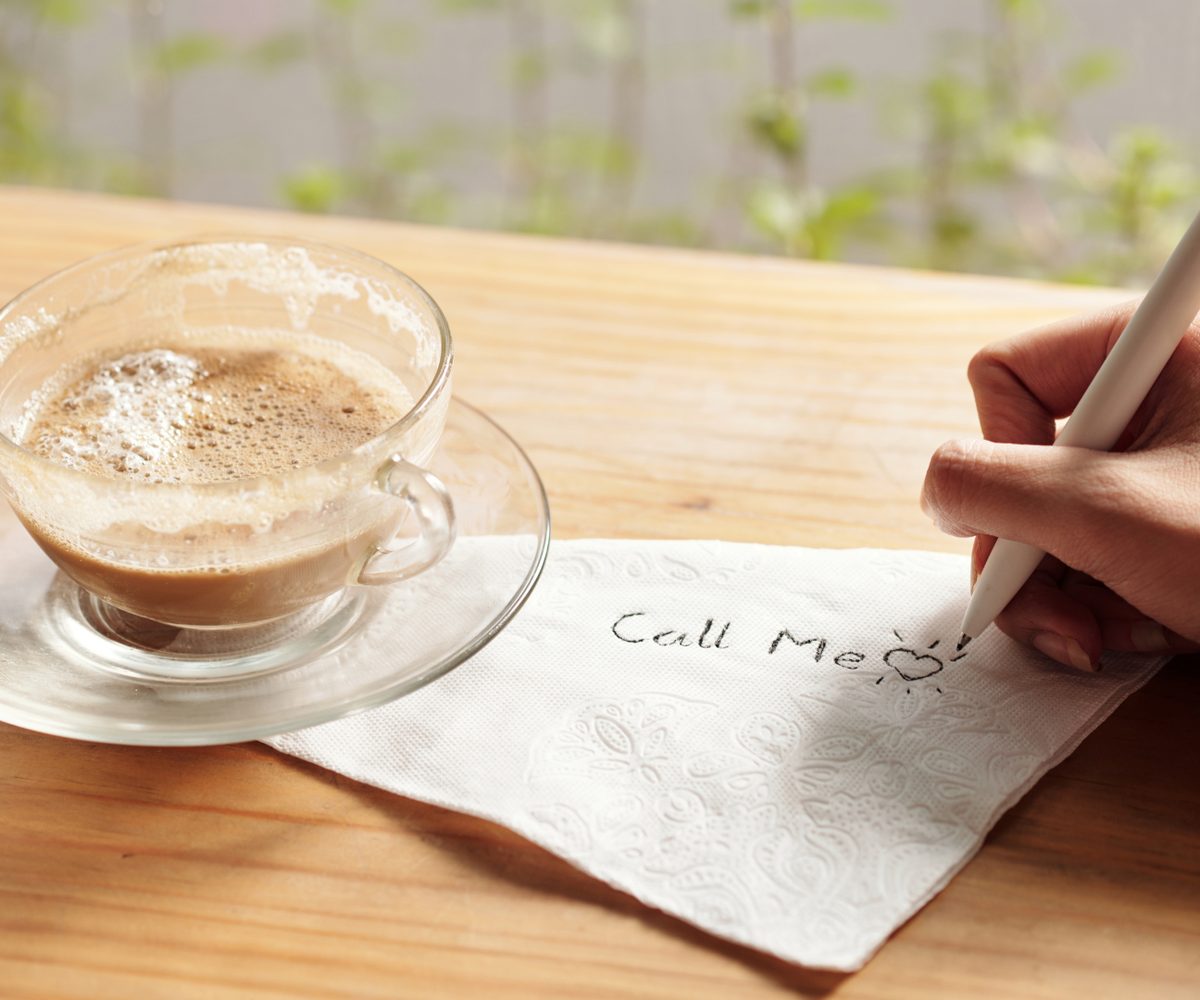 A glass cup of coffee on a wooden counter beside a napkin with "Call Me" written on it