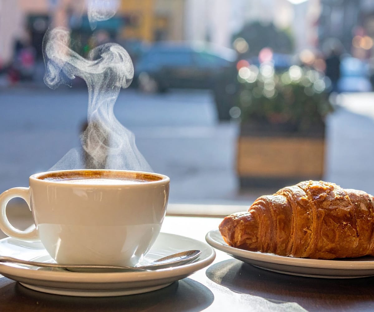 A cup of coffee and a croissant on a cafe table by a window