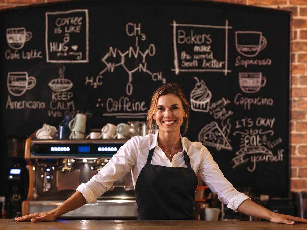 A barista behind the counter, a chalkboard of drinks on the wall behind her